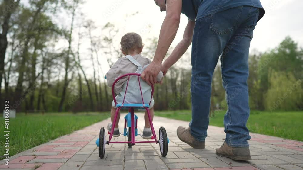 Father guides toddler riding tricycle outside in park. Family enjoys playful time together as father helps toddler learn to ride tricycle. Father supports child riding outside. Father outside park fun Father guides toddler riding tricycle outside in park. Family enjoys playful time together as father helps toddler learn to ride tricycle. Father supports child riding outside. Father outside park fun