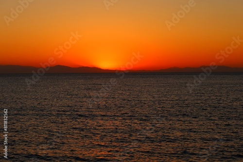 Calm morning sea, hills in the distance and red orange rising sun. Dawn by the sea, peaceful landscape travel photography. Distant land over the sea and colorful sunrise.