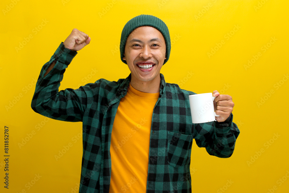 An excited young Asian man, dressed in a beanie hat and casual shirt, is enthusiastically making a strong gesture by lifting his arm and flexing his muscles while holding a morning coffee mug An excited young Asian man, dressed in a beanie hat and casual shirt, is enthusiastically making a strong gesture by lifting his arm and flexing his muscles while holding a morning coffee mug