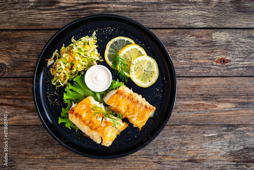 Fried codfish loin served with cabbage salad and lemon slices on black plate on wooden table
