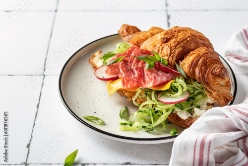 Croissant sandwich with salami sausage, cheese and cucumber in a plate for tasty breakfast on white tile table, text space