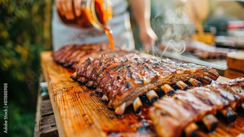 Grilled ribs are laid on a wooden cutting board, and in the backdrop is a blurry high angle shot of an unnamed cook pouring barbecue