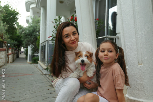 Mom, daughter and a wire haired jack russell terrier pup hugging on the porch of their house. Close up, copy space, background.