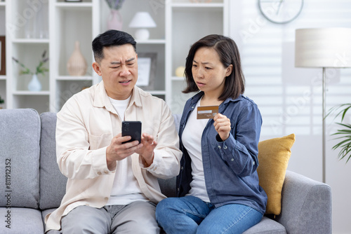 Canvas-taulu Asian couple looks distressed as they uncover unauthorized charges on their credit card phone, man confusion while the woman, holding the card, seems anxious