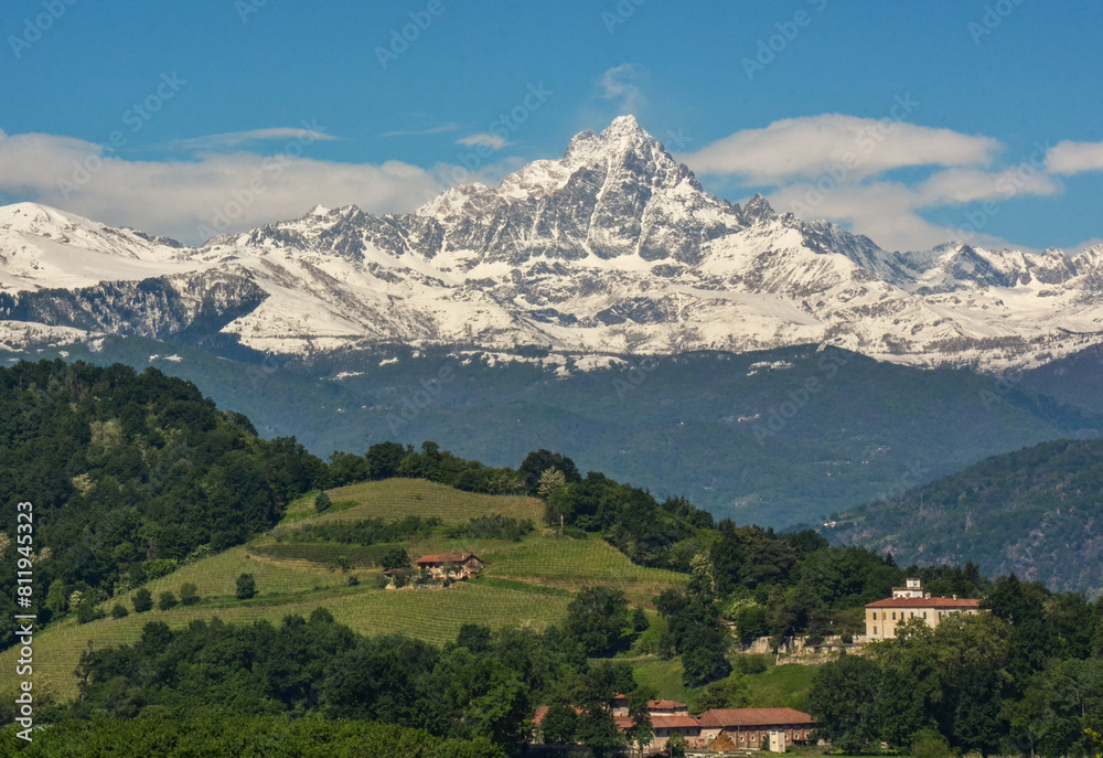 Monviso mountain range completely covered in snow in contrast with the green hills at the bottom of the valley on a spring day. Saluzzo, Monviso Natural Park, Piedmont, Italy.