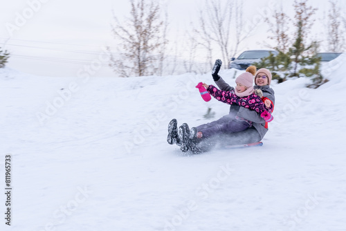 Two happy mother and daughter skating down a snowy hill on a sled. Two sisters in winter clothes have a rest in winter. Winter outdoor recreation. Family winter holidays