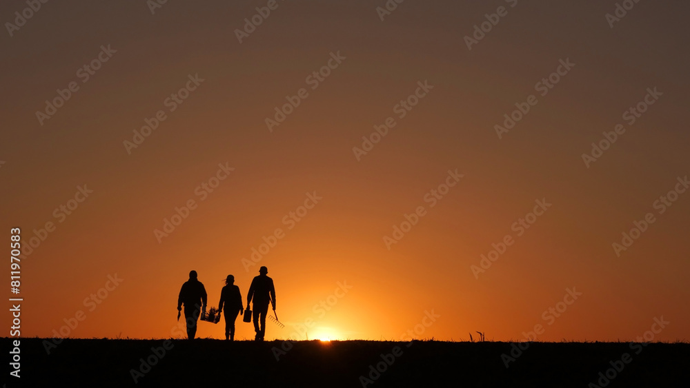 At sunset, three farmers walk through a field, carrying their equipment. At sunset, three farmers walk through a field, carrying their equipment.
