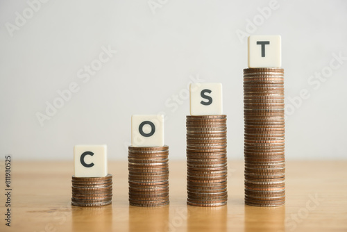 Obraz na plátně Word COST and stack coins as increase graph on wooden table with white wall background copy space
