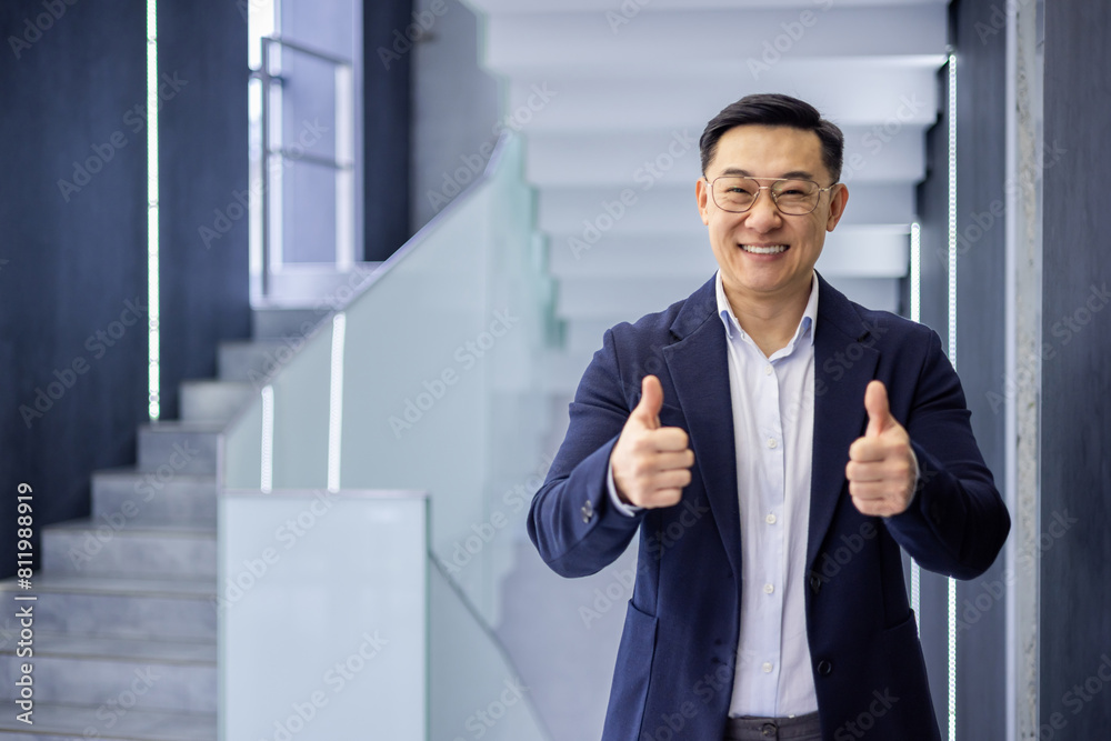 Confident Asian businessman standing by stairs in an office, giving thumbs up with a smile, promoting motivation and success. Confident Asian businessman standing by stairs in an office, giving thumbs up with a smile, promoting motivation and success.