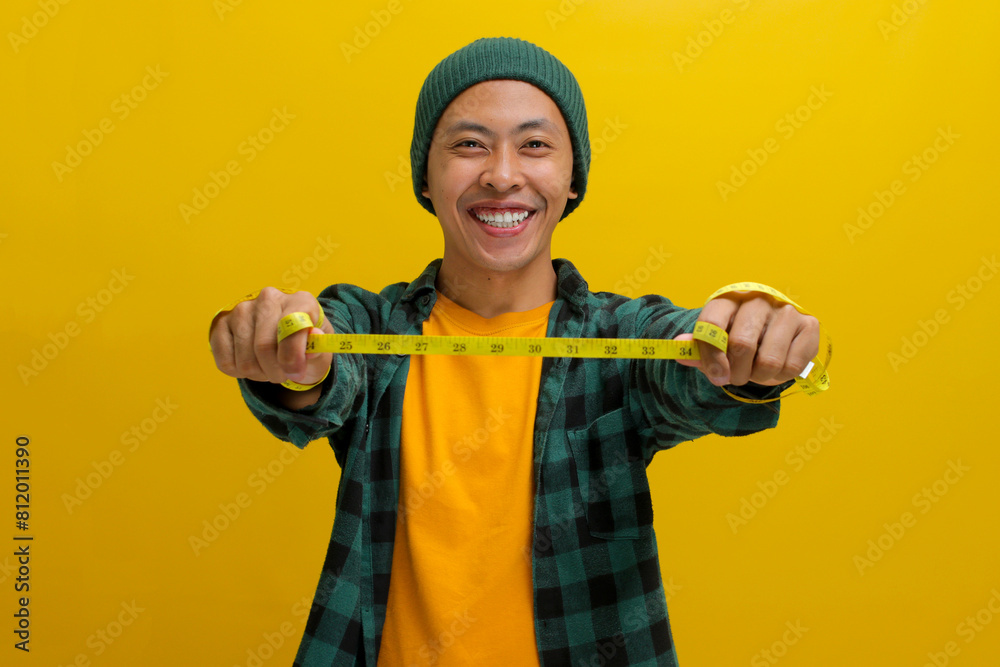 Cheerful Asian man in a beanie and casual clothes stretches a yellow measuring tape in front of him, seemingly measuring something. Isolated on a yellow background.