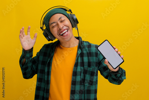 Asian man in a beanie and casual clothes listens to music on his headphones, phone held in hand. Isolated on a yellow background. photo