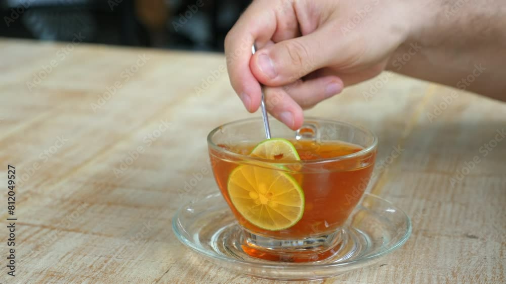 Person stirring fresh herbal tea with lemon in transparent cup on glass saucer, concept of natural remedies and healthy beverages. Person stirring fresh herbal tea with lemon in transparent cup on glass saucer, concept of natural remedies and healthy beverages.