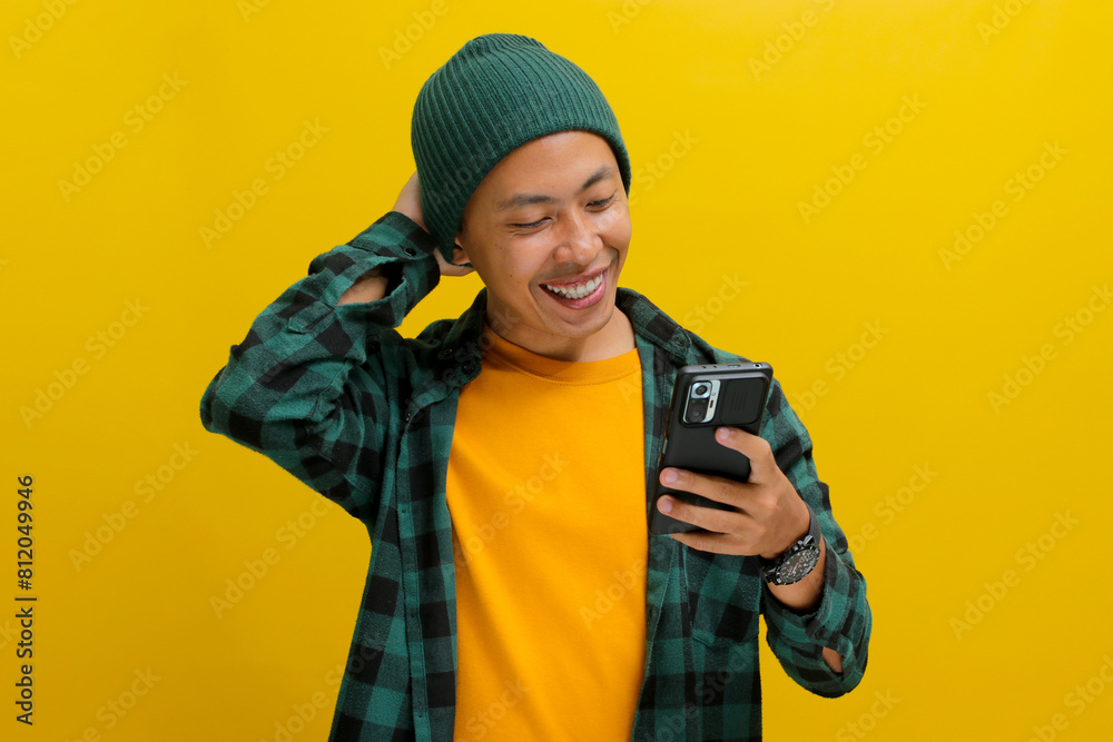 Happy young Asian man, wearing eyeglasses, a beanie hat, and casual shirt, reads a message on his mobile phone, visibly receiving good news while standing against a yellow background Happy young Asian man, wearing eyeglasses, a beanie hat, and casual shirt, reads a message on his mobile phone, visibly receiving good news while standing against a yellow background