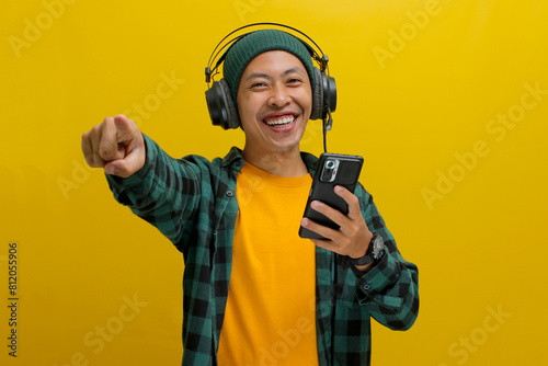 Asian man in a beanie and casual clothes points his finger directly at the camera while listening to music on his headphones. His phone is in his hand. Isolated on a yellow background.