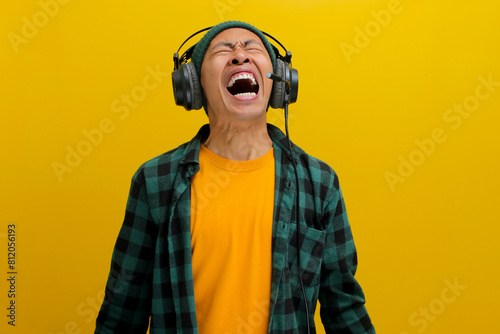 Annoyed Asian man in a beanie and casual clothes groans with frustration while wearing headphones. Loud music or noise is implied. Isolated on a yellow background. © Jamaludinyusup