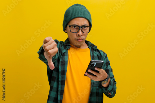A smart young Asian man, wearing eyeglasses and dressed in a beanie hat and casual shirt gives a thumbs-down gesture while holding a phone, expressing disapproval, disagreement and giving a bad review
