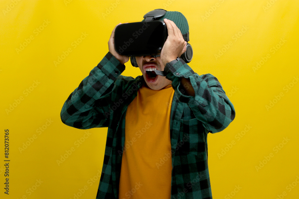 Terrified Asian man wearing a VR headset screams in fear while experiencing a horror movie in VR. Isolated on a yellow background Terrified Asian man wearing a VR headset screams in fear while experiencing a horror movie in VR. Isolated on a yellow background
