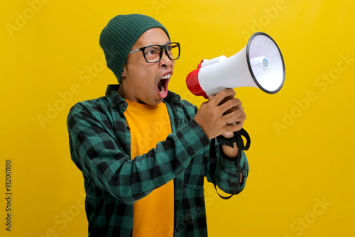 Young Asian man in a beanie hat and casual shirt shouts through a megaphone, expressing annoyance and frustration with angry yelling. Isolated on a yellow background photo