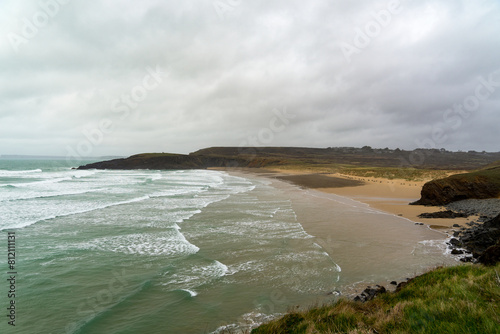 Vue sur la plage de Lostmarc'h, encadrée par des falaises et un éperon rocheux, avec la mer d'Iroise aux couleurs émeraude et écume blanche, sous un ciel couvert, un spectacle naturel saisissant. photo