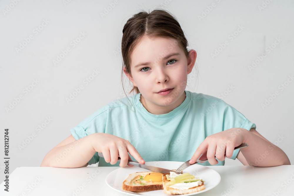 A cute little girl is having breakfast. A healthy breakfast of eggs, toast, cottage cheese and cucumber on her plate. Close-up. Selected focus A cute little girl is having breakfast. A healthy breakfast of eggs, toast, cottage cheese and cucumber on her plate. Close-up. Selected focus