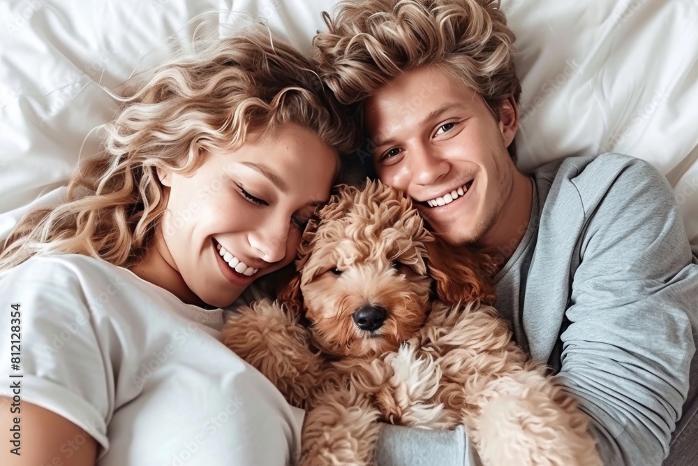 A smiling young woman and a guy hug a cute curly dog lying on a light bed A smiling young woman and a guy hug a cute curly dog lying on a light bed
