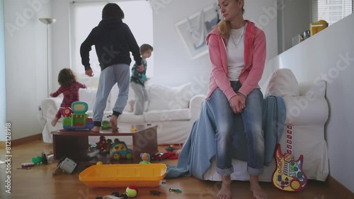 Exhausted mother sits with eyes closed and lying back on a couch, surrounded by her playful children and a disheveled living room strewn with toys, slow motion.
