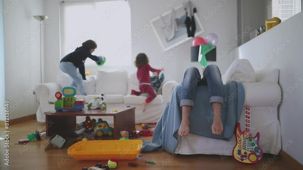 Exhausted mother sits with eyes closed and lying back on a couch, surrounded by her playful children and a disheveled living room strewn with toys, slow motion.