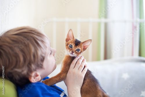 Happy child looking at abyssinian ruddy kitten in hands. Little boy playing with red kitten at home. Pets care.  Image for websites about cats. Selective focus.