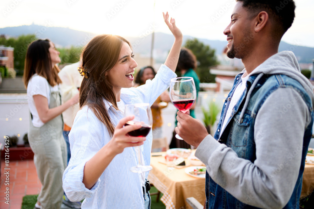 © CarlosBarquero - Excited multiracial young couple celebrating rooftop party drinking red wine. Friends dancing at sunset date weekend gathering. Cheerful gen z people enjoying romantic leisure outdoors.