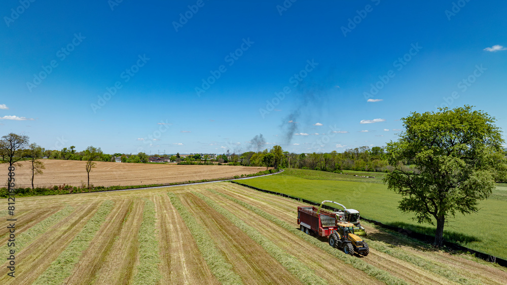 An expansive aerial photograph highlights the dynamic nature of farming with active harvesting machinery in the foreground, set against a backdrop of mixed greenery and brown fields. An expansive aerial photograph highlights the dynamic nature of farming with active harvesting machinery in the foreground, set against a backdrop of mixed greenery and brown fields.