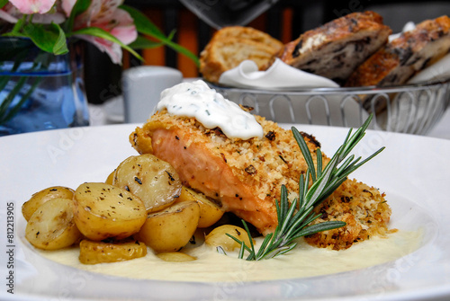 Salmon, breaded salmon fillet topped with sour cream bechamel sauce, roasted potatoes and slices of bread on restaurant table