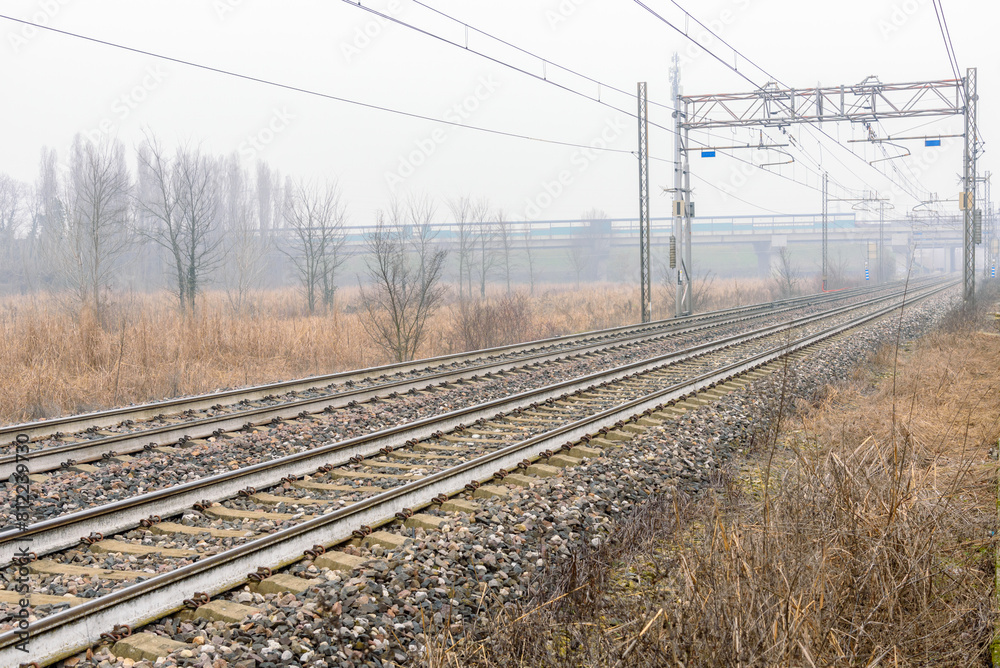 Deserted electric railway running under a motorway bridge in the countryside on a foggy winter morning