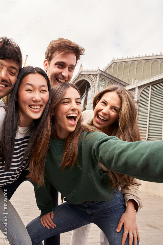 Fototapeta premium Vertical. Group happy young people smiling making gestures while taking selfie together at travel event. Generation z friends having fun and sharing positive moment posing cheerful for photo outdoors