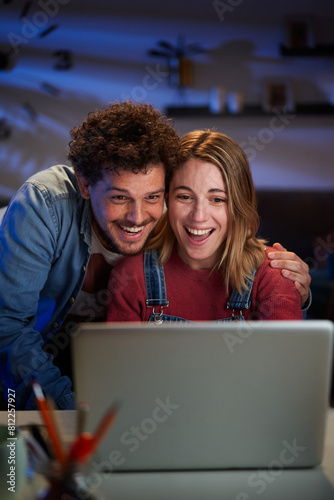 Vertical excited young adult Caucasian couple looking at laptop surprised celebrating successful start-up business. Millennial man and woman happy with positive results of online work at night home 