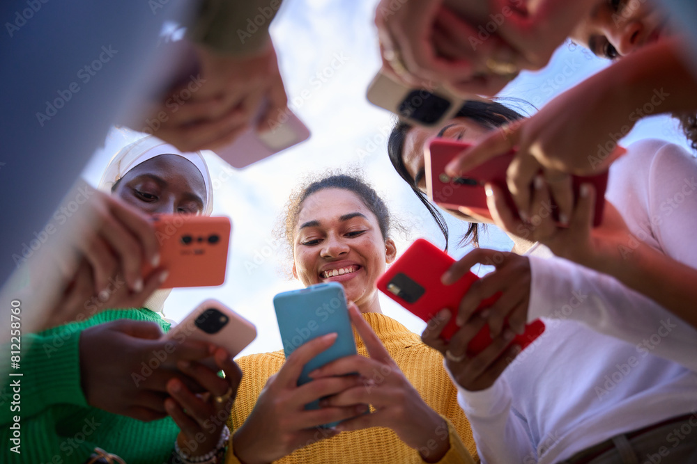 Obraz premium Low angle. Group of multiracial young women smiling gathered in circle using mobile phones. Cheerful females looking cells concentrated. Addiction to technology of gen Z