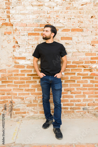 Confident young hispanic man wearing a blank black t-shirt for mockup presentation, standing against a rustic brick wall background © AntonioDiaz