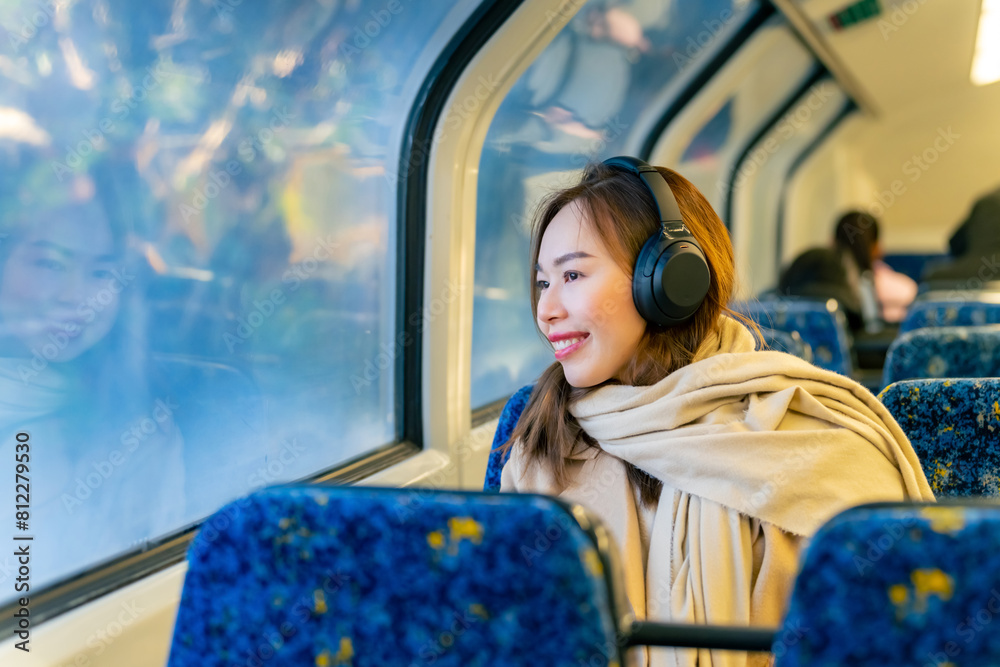 Fototapeta premium Asian woman using mobile phone and listening to music or podcast on headphones during travel on train in the city. Attractive girl enjoy urban lifestyle with digital gadget device and online network.