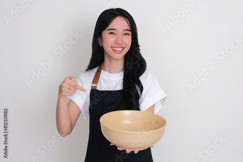 Asian woman wearing apron smiling and pointing to empty serving bowl that she hold photo