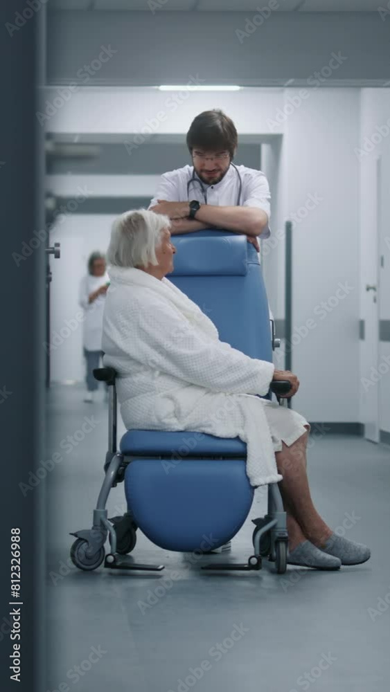 Male doctor stands with female patient in wheelchair in clinic corridor. Medic talks to elderly woman before surgery near operating room. Medical staff, patients in hospital or medical center hallway. Male doctor stands with female patient in wheelchair in clinic corridor. Medic talks to elderly woman before surgery near operating room. Medical staff, patients in hospital or medical center hallway.