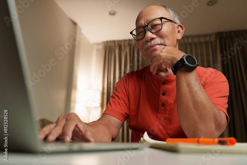 Confident Asian senior man using a laptop at home, deep in thought and planning