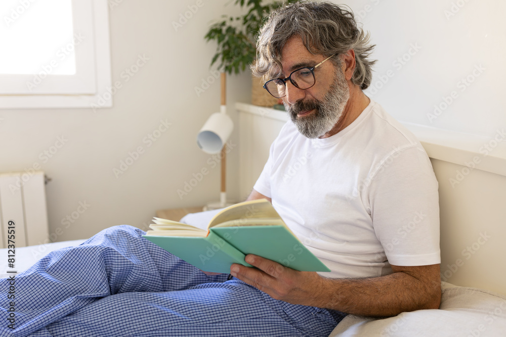 Mature man with glasses relaxing in bed, reading a book wearing pyjamas.