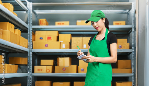 Asian woman servicer checking stock of package in storeage room with note in apron wear for business