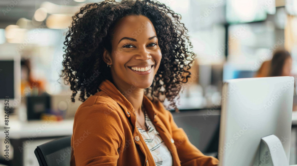 Portrait of a young African American woman working at a computer in a modern office. Smiling woman in office clothes looking at camera indoors. Portrait of a young African American woman working at a computer in a modern office. Smiling woman in office clothes looking at camera indoors.