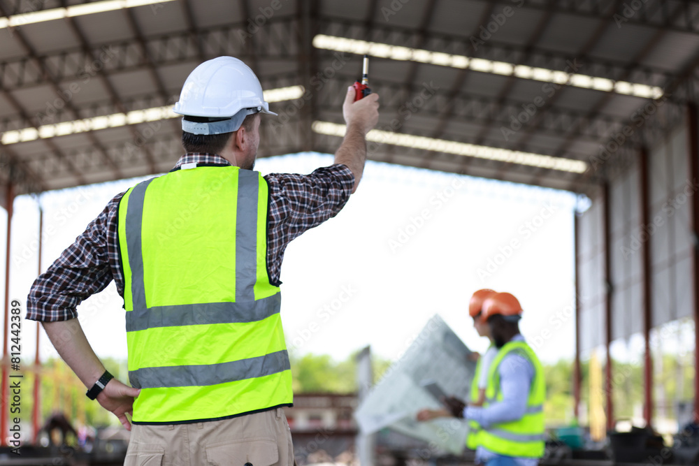 A senior civil engineer is seen from behind while working at a construction site. They are holding a walkie-talkie and overseeing the work progress. A senior civil engineer is seen from behind while working at a construction site. They are holding a walkie-talkie and overseeing the work progress.
