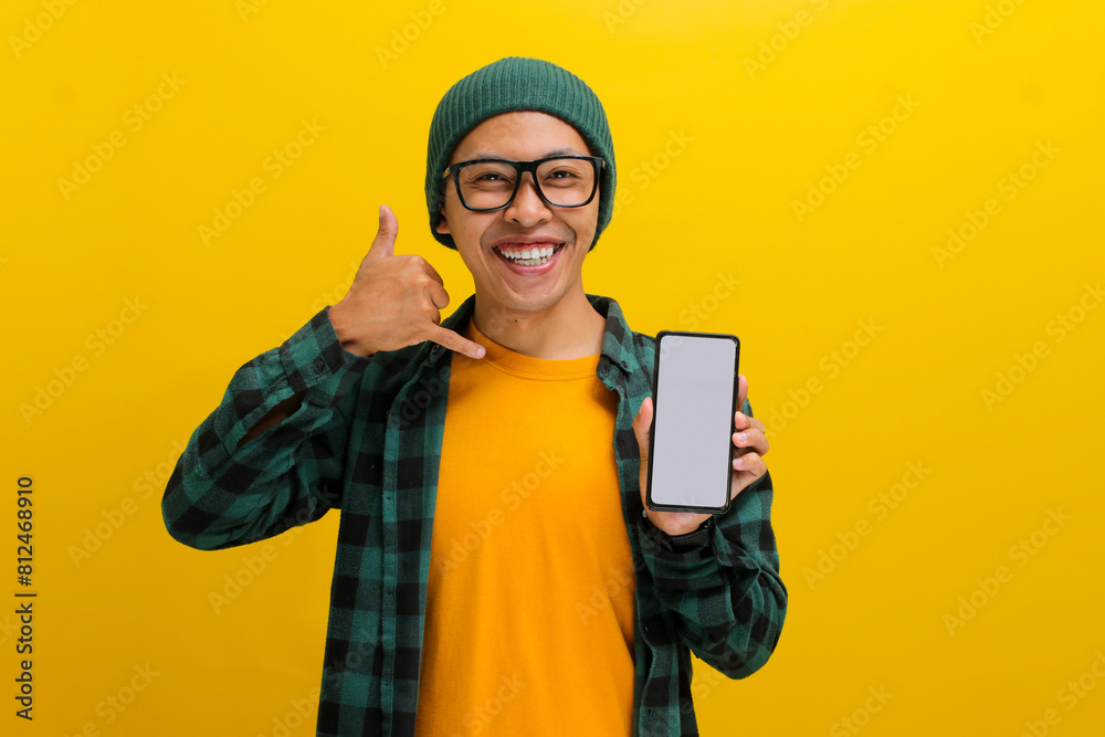 Cheerful Asian man in a beanie and casual shirt holds up his phone, screen facing the camera. He gestures a call me sign. Isolated on a yellow background. Communication, invitations, and contact. Cheerful Asian man in a beanie and casual shirt holds up his phone, screen facing the camera. He gestures a call me sign. Isolated on a yellow background. Communication, invitations, and contact.