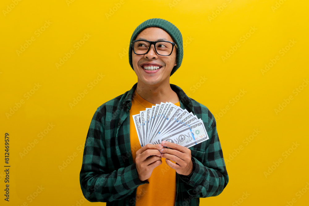 Daydreaming Asian man contemplates his vacation plans and investment options, holding cash thoughtfully while gazing upwards. Isolated on a yellow background. Financial planning, travel dreams concept Daydreaming Asian man contemplates his vacation plans and investment options, holding cash thoughtfully while gazing upwards. Isolated on a yellow background. Financial planning, travel dreams concept