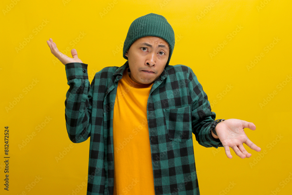 Confused young Asian man, dressed in a beanie hat and casual shirt is shrugging his shoulders in a gesture of cluelessness while standing against yellow background. Confused young Asian man, dressed in a beanie hat and casual shirt is shrugging his shoulders in a gesture of cluelessness while standing against yellow background.