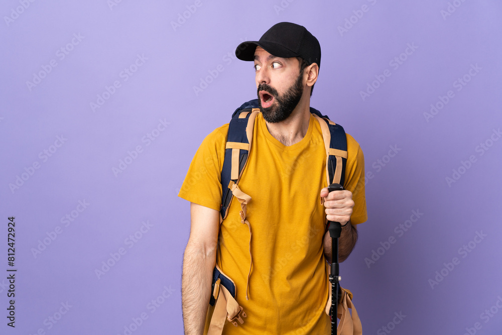 Caucasian handsome man with backpack and trekking poles over isolated background doing surprise gesture while looking to the side Caucasian handsome man with backpack and trekking poles over isolated background doing surprise gesture while looking to the side