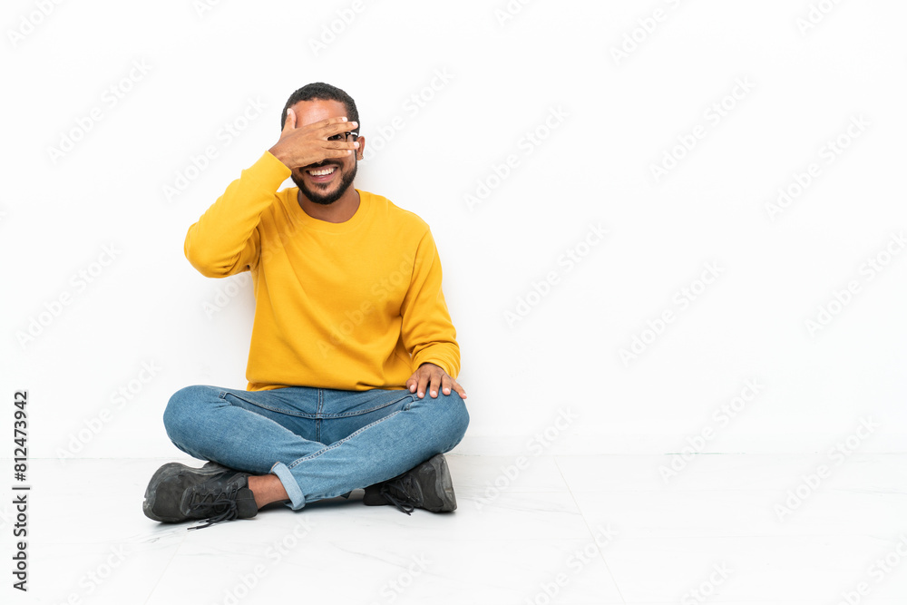 Young Ecuadorian man sitting on the floor isolated on white wall covering eyes by hands and smiling