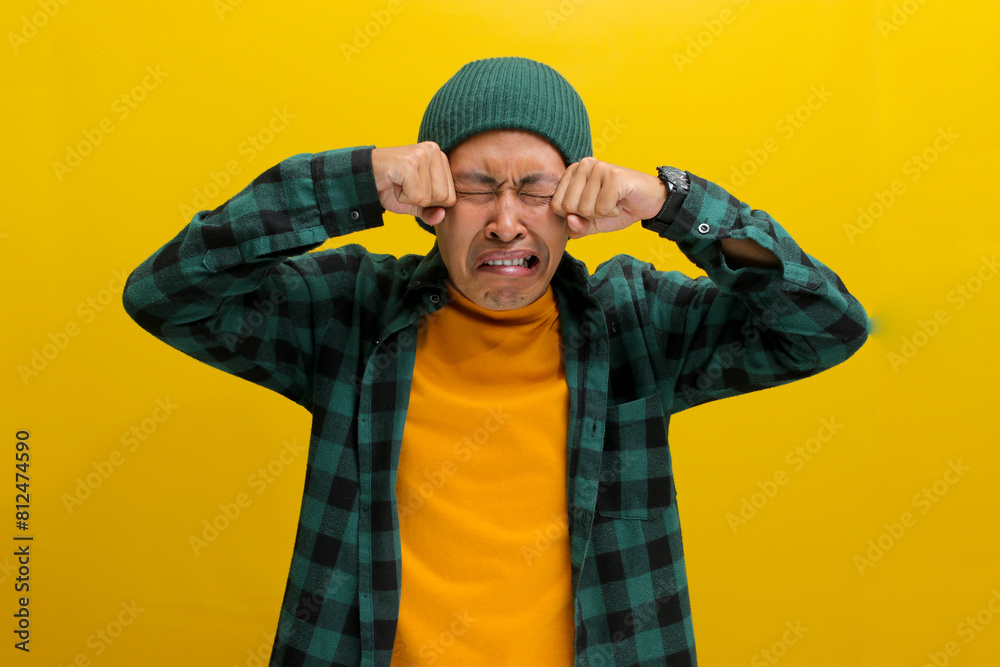 Distressed young Asian man, dressed in a beanie hat and casual shirt, rubs his eyes, tears streaming down his face, expressing deep upset and unhappiness while standing against a yellow background Distressed young Asian man, dressed in a beanie hat and casual shirt, rubs his eyes, tears streaming down his face, expressing deep upset and unhappiness while standing against a yellow background